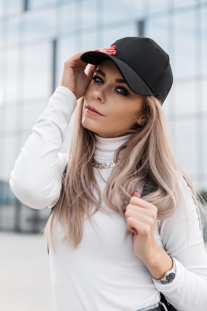 Retrato urbano de una hermosa joven con una gorra negra de moda en un suéter blanco con una cadena caminando por la ciudad