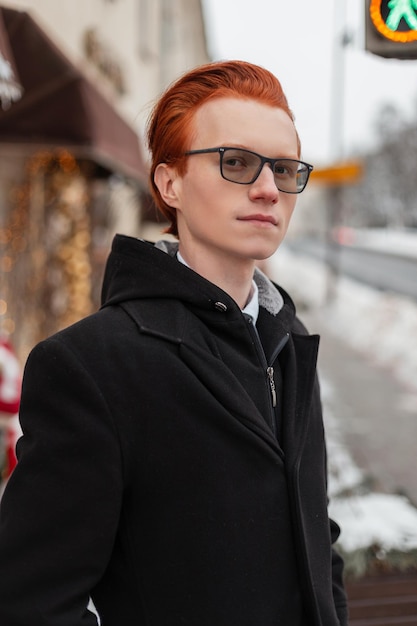 Retrato urbano de un hermoso joven pelirrojo con anteojos en ropa de negocios de moda con abrigo, camisa y corbata camina por la calle.