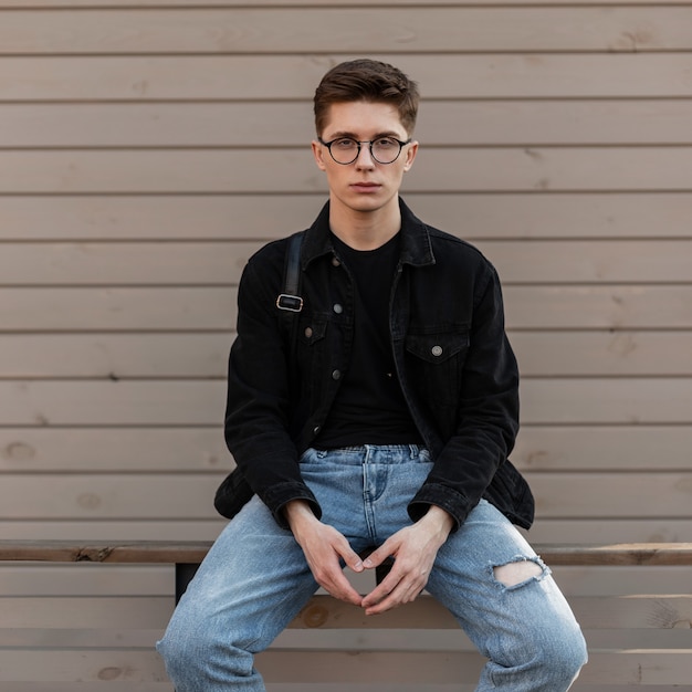 Retrato urbano jovem sério com óculos vintage em jeans e mochila na rua