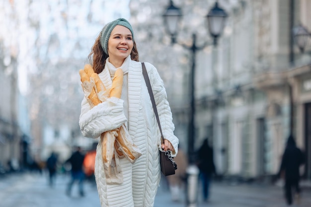 Retrato urbano de una mujer joven caminando por la calle sosteniendo unos panes y sosteniendo una bolsa en su hombro. Regreso de la tienda. Una sonrisa luminosa y radiante. El concepto de panadería.