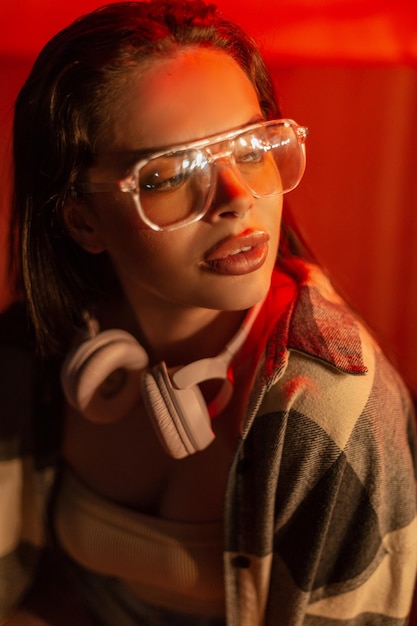 Retrato urbano nocturno de una hermosa mujer de moda con gafas de moda y auriculares se sienta en la ciudad nocturna cerca de la luz roja