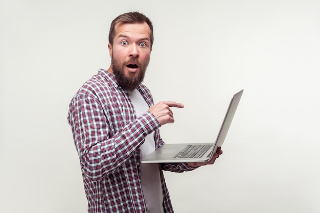 Retrato de un usuario de computadora conmocionado hombre barbudo en camisa a cuadros casual apuntando a la computadora portátil que parece sorprendido por la aplicación de computadora increíble noticias de Internet tiro de estudio interior fondo blanco