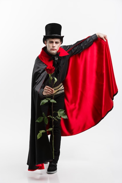 Foto retrato del vampiro caucásico hermoso que sostiene la rosa hermosa roja en el fondo blanco del estudio.