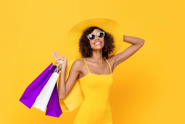 Retrato de verano de una alegre y sonriente mujer afroamericana con gafas de sol sosteniendo su sombrero y bolsas de compras en un estudio aislado de fondo amarillo