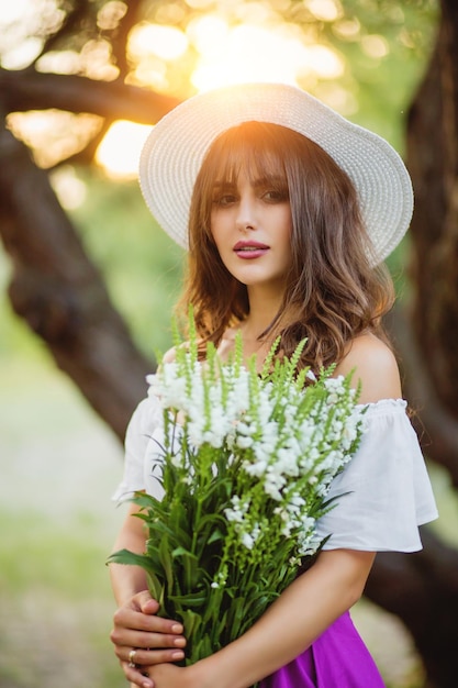 Retrato de verano hermosa joven con un sombrero blanco al atardecer en el parque