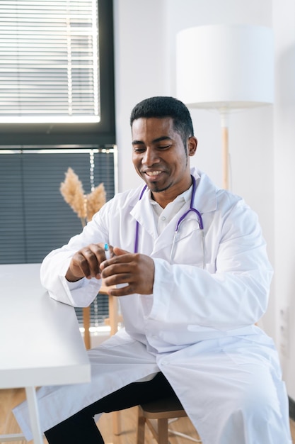 Retrato vertical de un alegre médico afroamericano con uniforme blanco sosteniendo una jeringa en la mano sentado en la mesa en una clínica médica
