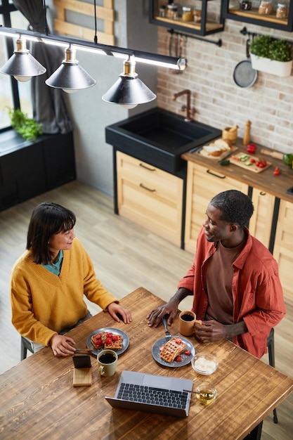 Retrato vertical de alto ángulo de una joven pareja de razas mixtas disfrutando del desayuno en un acogedor espacio de copia de cocina casera