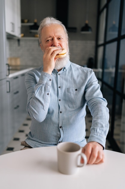 Retrato vertical de un anciano maduro hambriento comiendo comida rápida disfrutando de la cena en casa y bebiendo café