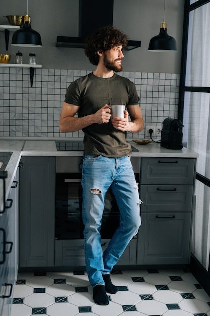 Retrato vertical de un apuesto joven pensativo sosteniendo una taza en la mano con café matutino mirando hacia otro lado de pie en la cocina