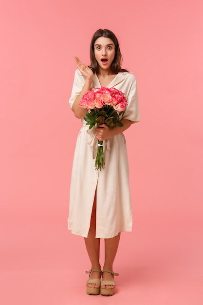 Retrato vertical de cuerpo entero, la niña bastante sorprendida recibió un regalo inesperado, recibió la entrega sosteniendo flores, jadeando con la boca abierta asombrada y sin palabras con expresión atónita, rosa