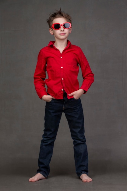 Retrato vertical de cuerpo entero de un niño elegante con gafas de sol rojas sobre un fondo gris