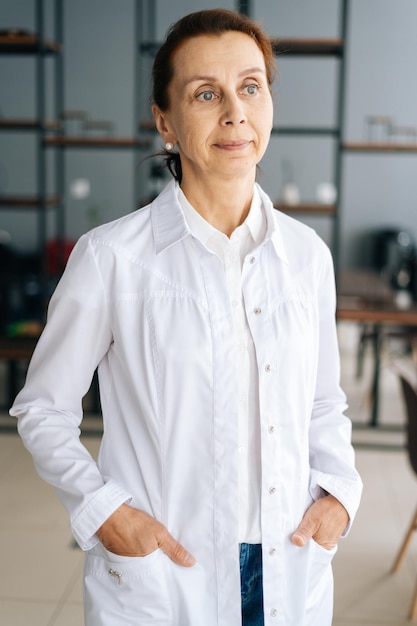 Retrato vertical da médica pensativa em uniforme médico branco em pé no escritório do hospital, olhando para longe. Vista frontal da mulher praticante atraente bem sucedida posando no local de trabalho.