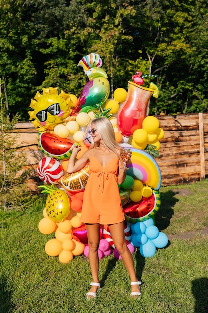 Retrato vertical de bela loira bronzeada em vestido de verão e óculos de sol posando com vários balões de hélio festivos coloridos em um dia ensolarado