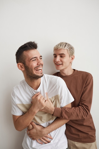 Foto retrato vertical de casal gay despreocupado se abraçando e rindo alegremente enquanto posa contra um branco em casa