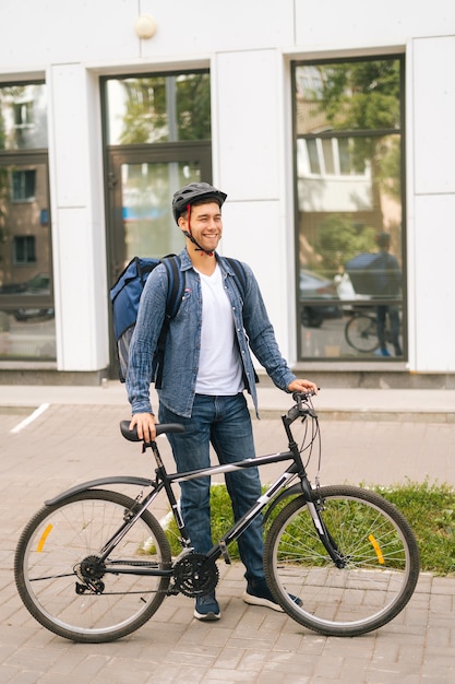 Retrato vertical de comprimento total de rir bonito jovem entregador de capacete posando em pé perto de uma bicicleta na rua da cidade, olhando para longe. Mensageiro masculino com mochila térmica entregando comida ao cliente