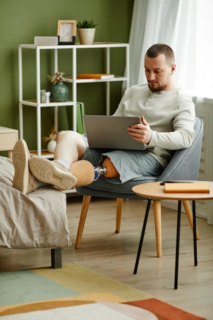 Retrato vertical de corpo inteiro de homem adulto com perna protética relaxando em casa e usando laptop