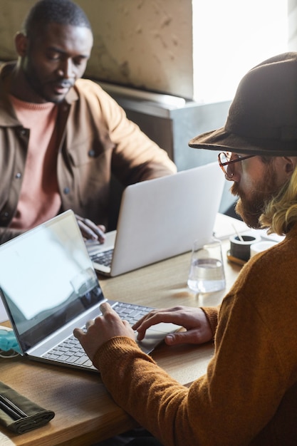 Retrato vertical de dois homens contemporâneos usando laptops enquanto trabalhava em uma mesa de café iluminada pela luz do sol no interior do loft