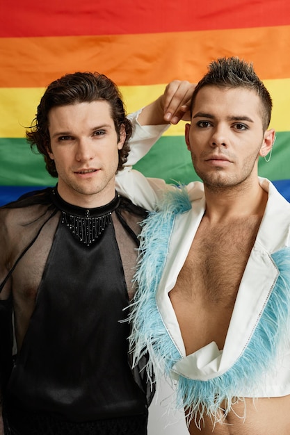 Foto retrato vertical de dois jovens gays posando com a bandeira do orgulho lgbtq ao fundo e usando extrav