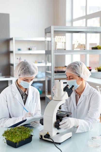 Retrato vertical de duas jovens cientistas olhando no microscópio enquanto estudam amostras de plantas no laboratório de biotecnologia