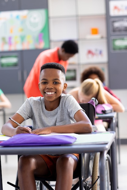 Retrato vertical de estudante afro-americano sorridente em cadeira de rodas na mesa no espaço de cópia da classe