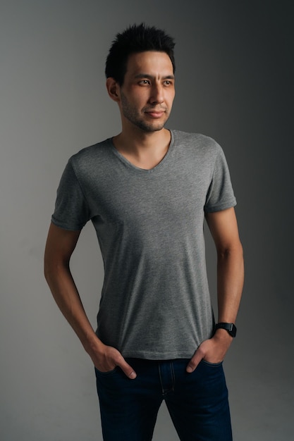 Retrato vertical de jovem confiante em pé de t-shirt cinza com as mãos nos bolsos no fundo escuro isolado. Foto de estúdio de homem caucasiano vestindo roupas casuais posando olhando para longe