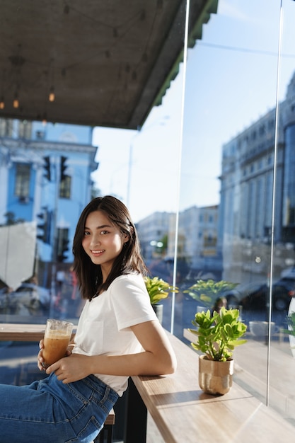 Retrato vertical de jovem modelo feminino asiático bebendo café em um café perto da janela