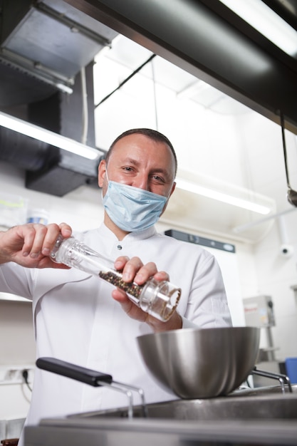 Foto retrato vertical de um chef em máscara facial médica salgando refeições preparadas na cozinha do restaurante