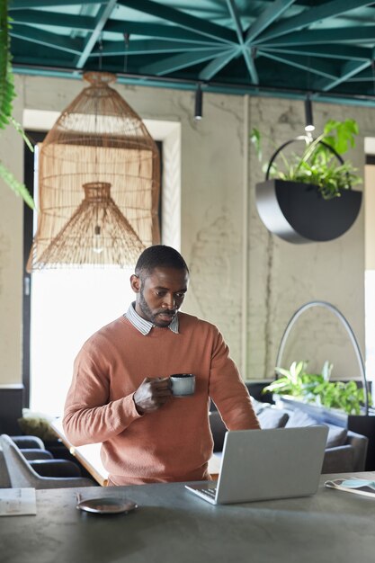 Retrato vertical de um homem afro-americano usando um laptop e tomando café enquanto trabalha em um café ecológico decorado com plantas verdes frescas