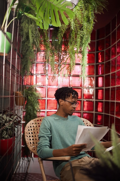 Retrato vertical de um homem contemporâneo africano trabalhando com documentos enquanto está sentado em um café decorado com plantas