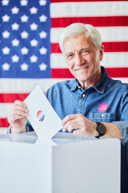 Foto retrato vertical de um homem sênior sorridente colocando cédula na lixeira contra a bandeira americana no fundo