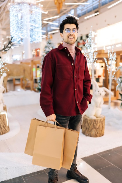 Retrato vertical de um jovem barbudo feliz em copos segurando sacos de papel com presentes sorrindo olhando para a câmera em pé no corredor do shopping center