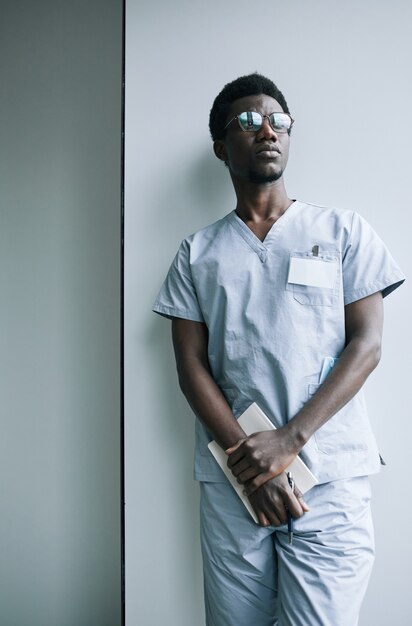 Foto retrato vertical de um jovem médico afro-americano parado perto da parede na clínica e olhando para longe