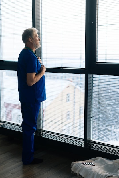Retrato vertical de um médico maduro pensativo olhando para fora pela janela Cirurgião sério concentrado em terno azul de praticante no hospital