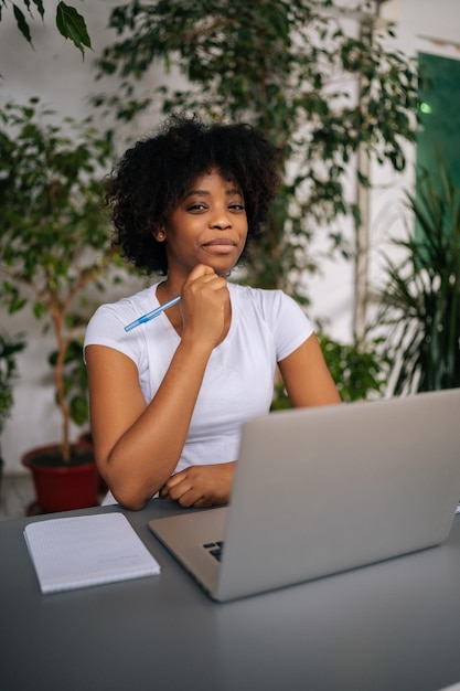 Retrato vertical de uma empresária afro-americana bem sucedida segurando a caneta na mão séria olhando para a câmera sentada à mesa na sala de escritório em casa leve