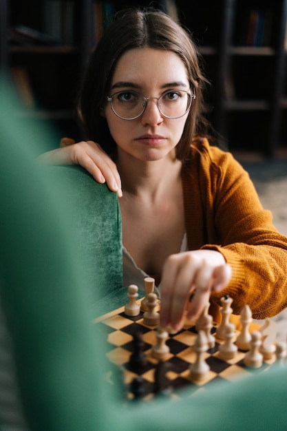 Retrato vertical de uma jovem atraente em óculos elegantes fazendo movimento de xadrez com peça de cavaleiro sentado no chão olhando para a câmera