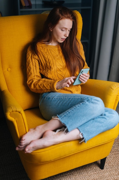 Retrato vertical de uma jovem bonita usando smartphone sentado em uma poltrona amarela olhando para a tela do dispositivo Linda mulher relaxando conversando no celular
