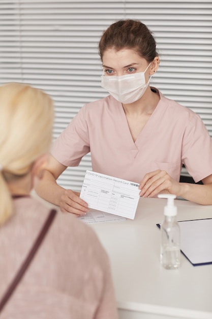 Retrato vertical de uma jovem enfermeira usando máscara enquanto registra pacientes para vacinação secreta no centro médico