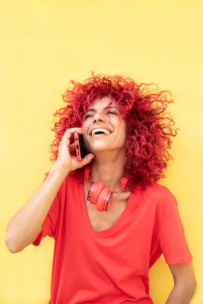 Foto retrato vertical de uma jovem latina com cabelo afro vermelho falando no celular e rindo encostado na parede amarela fones de ouvido vermelhos no pescoço