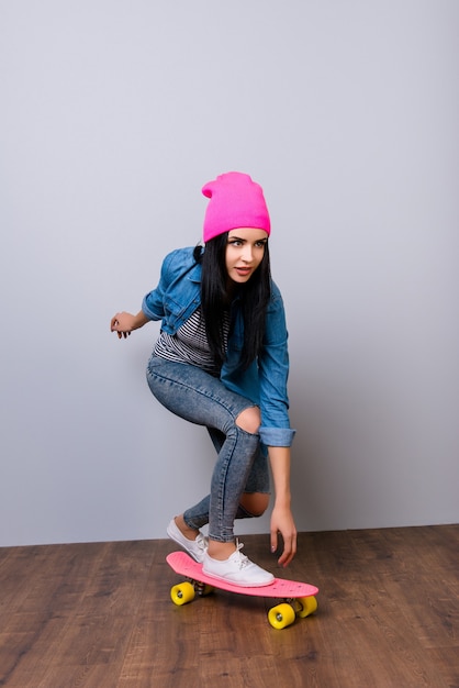 Retrato vertical de uma jovem mulher bonita em jeans e chapéu rosa andando de skate contra o espaço cinza