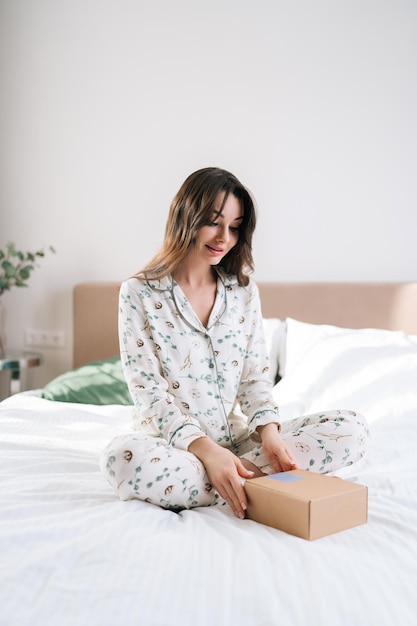 Retrato vertical de uma jovem romântica feliz vestindo pijama sentada na cama com uma caixa de presente surpresa embrulhada no dia dos namorados festivo