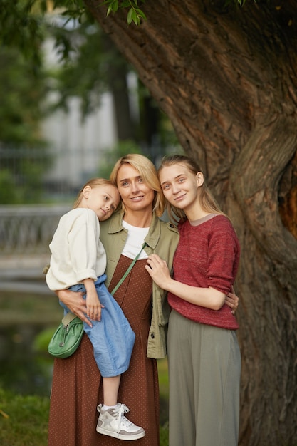 Retrato vertical de uma mãe adulta moderna com duas filhas posando juntas sorrindo alegremente ao lado de uma árvore ao ar livre, aproveitando o tempo com a família no parque