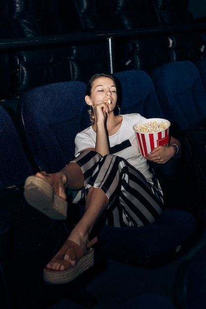 Retrato vertical de uma mulher sentada em uma cadeira em um cinema comendo uma tigela de popcorn