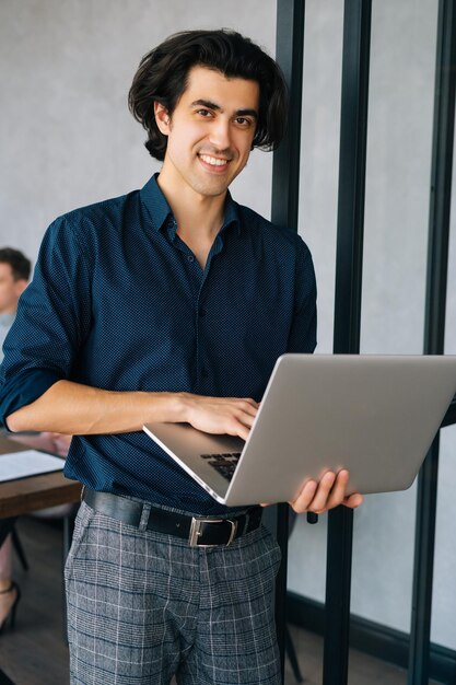 Retrato vertical do empresário alegre em roupas casuais, segurando o laptop na mão, em pé na sala do escritório, olhando para a equipe de negócios de inicialização da câmera discutindo o projeto sentado na mesa no fundo