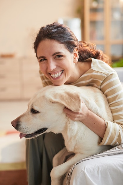 Foto retrato vertical em tons quentes de uma jovem sorridente abraçando o cachorro sentado na cama no interior de uma casa aconchegante