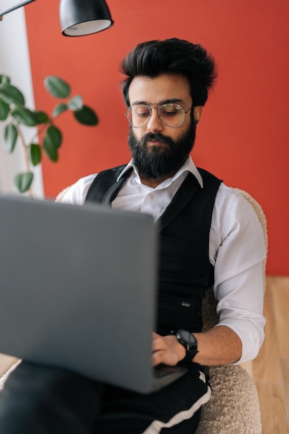 Retrato vertical de un exitoso hombre de negocios indio con elegante ropa formal que trabaja en una laptop sentada en una silla en una oficina de trabajo ligera