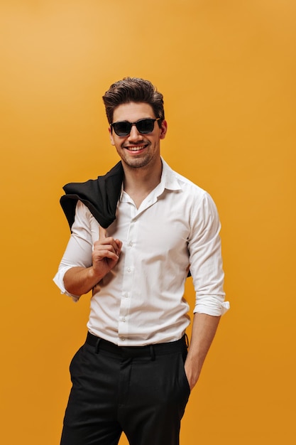 Retrato vertical de un hombre brunet guapo y feliz con camisa blanca, pantalones negros y gafas de sol sosteniendo una chaqueta y sonriendo con un fondo naranja