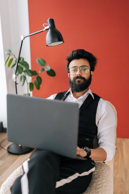 Retrato vertical de un hombre de negocios indio seguro de sí mismo con elegante ropa formal que trabaja en una laptop sentada en una silla en una oficina de trabajo ligera mirando la cámara