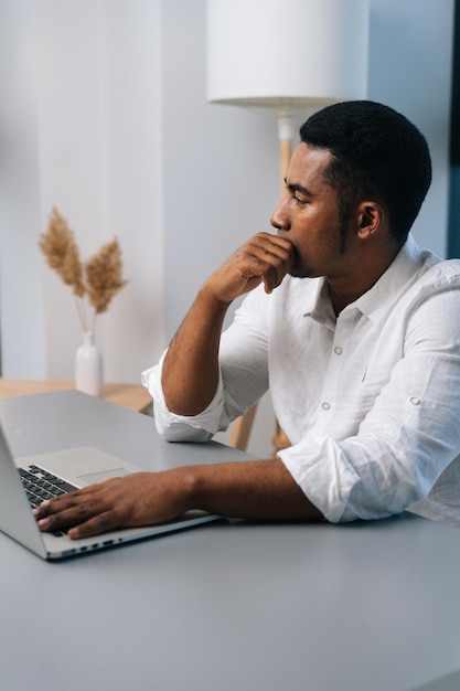Retrato vertical de un hombre de negocios negro pensativo que trabaja en una estrategia de negocios en una laptop sentada en el escritorio pensando en mirar hacia otro lado
