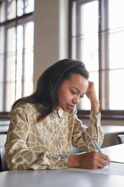 Retrato vertical de la joven afroamericana tomando el examen en la escuela mientras está sentado en un escritorio en la universidad y pensando, copie el espacio