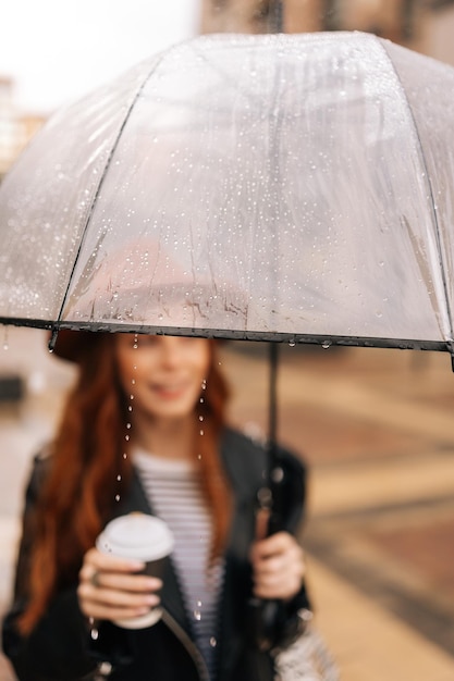 Retrato vertical de una joven alegre con sombrero de moda sosteniendo en la mano una taza con café caliente de pie con un paraguas transparente en la fría lluvia de otoño